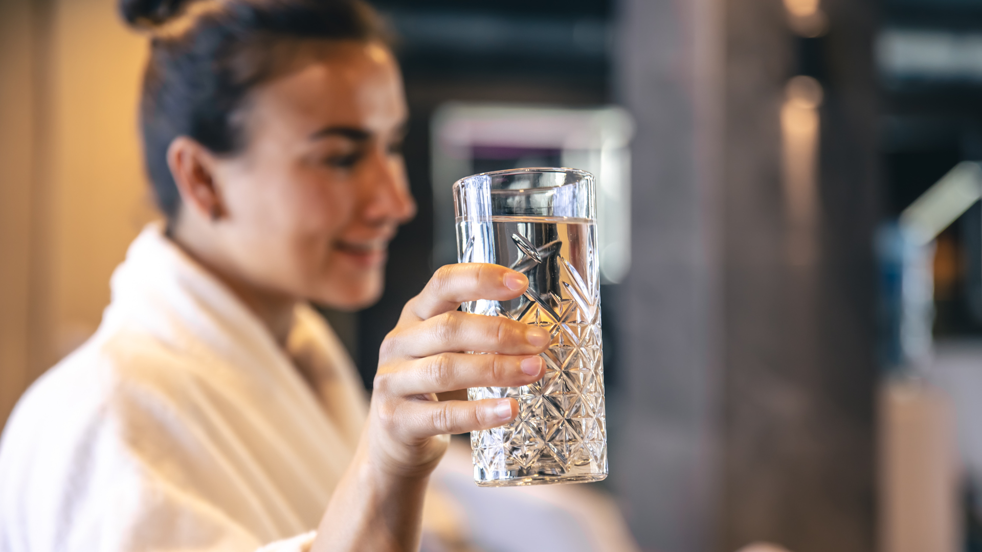 Eine Person trägt ein weißes Gewand und hält in einem sanft beleuchteten Innenraum ein dekoratives, mit Wasser gefülltes Glas.