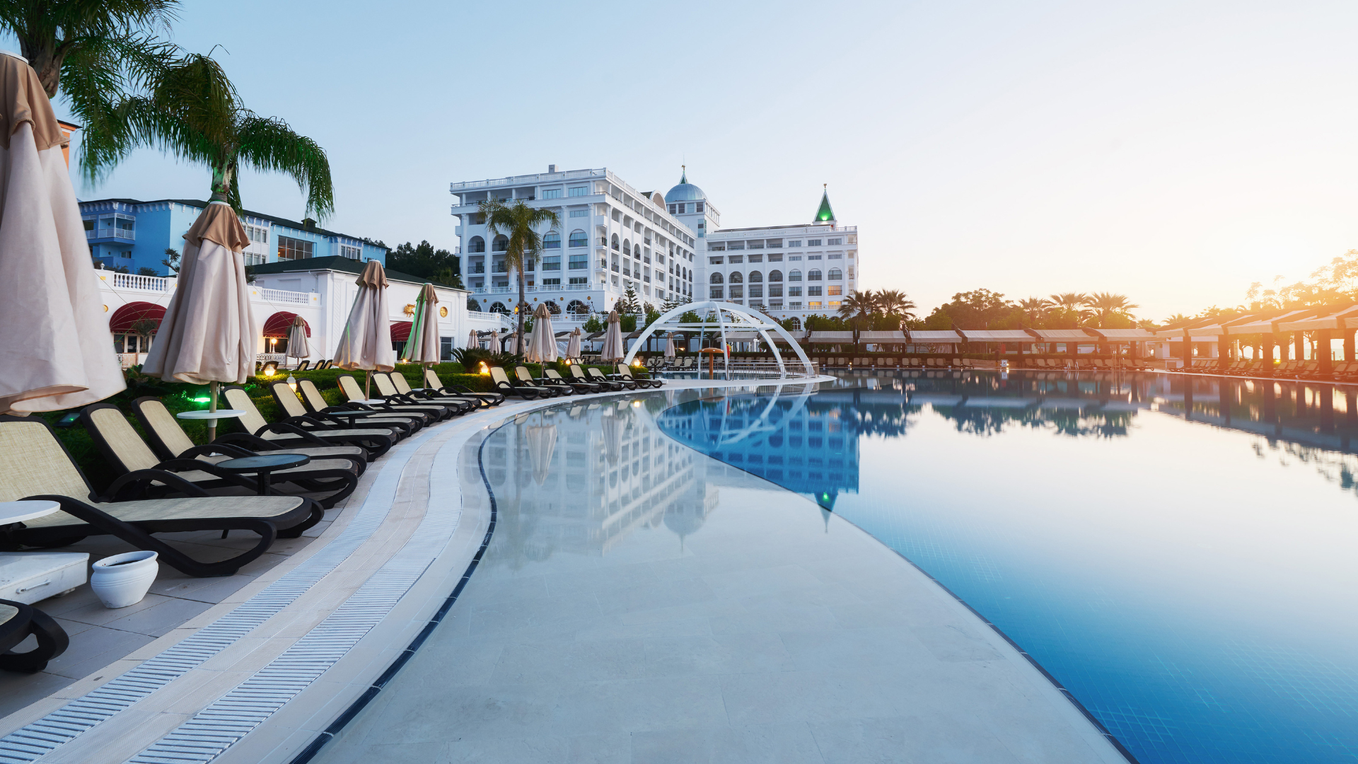 Ein großer, luxuriöser Pool mit Liegestühlen und Sonnenschirmen schmiegt sich um ein Deck. Im Hintergrund ist ein großes Hotelgebäude zu sehen, darüber Palmen und ein klarer blauer Himmel.