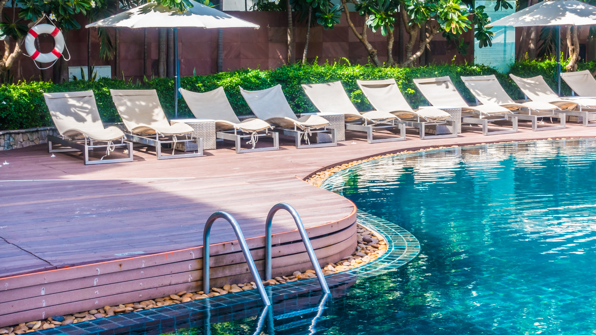 Szene am Pool mit einer Reihe leerer Liegestühle unter Sonnenschirmen, neben einem blauen Swimmingpool mit einer Leiter und umgebendem Grün.