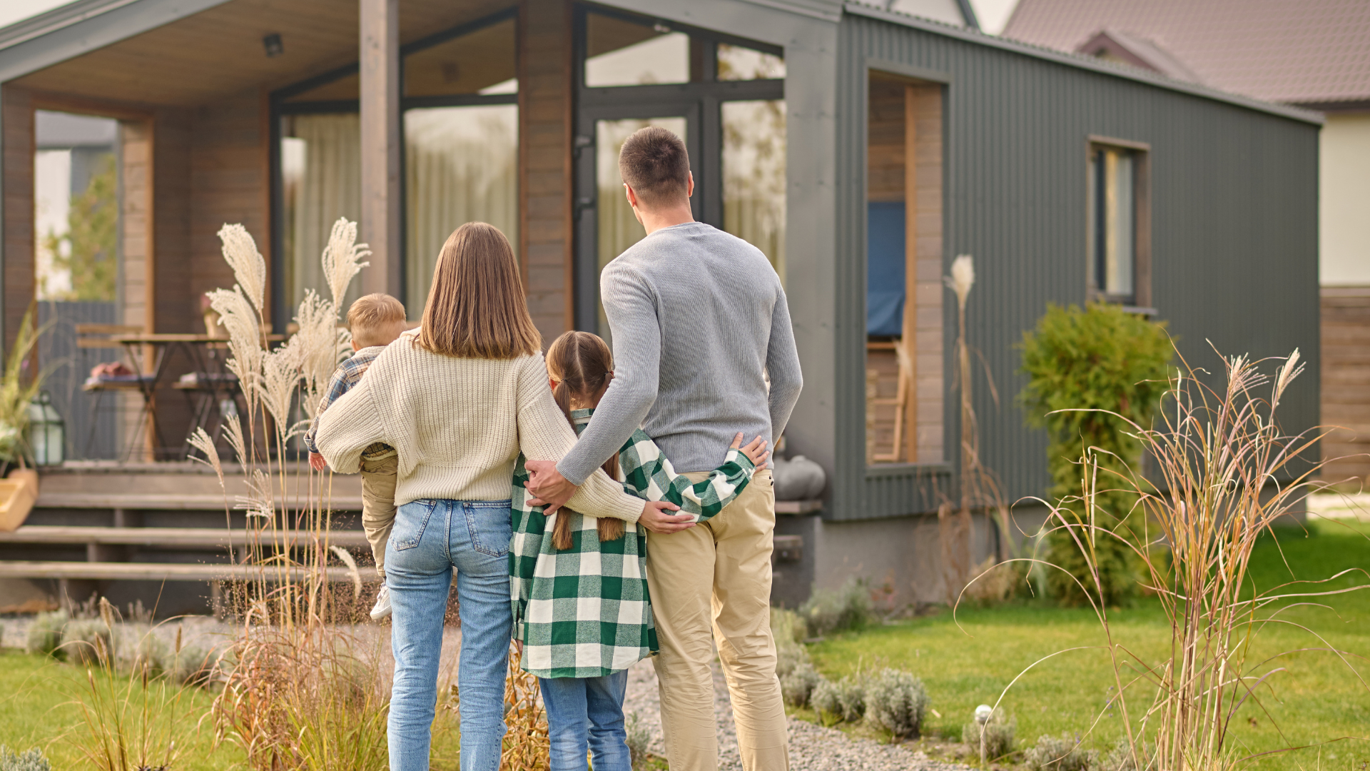 Eine vierköpfige Familie (von hinten zu sehen) steht in einem Garten vor einem modernen Haus. Die Erwachsenen halten ein Kind im Arm und umarmen ein älteres Kind, während sie zum Haus blicken.