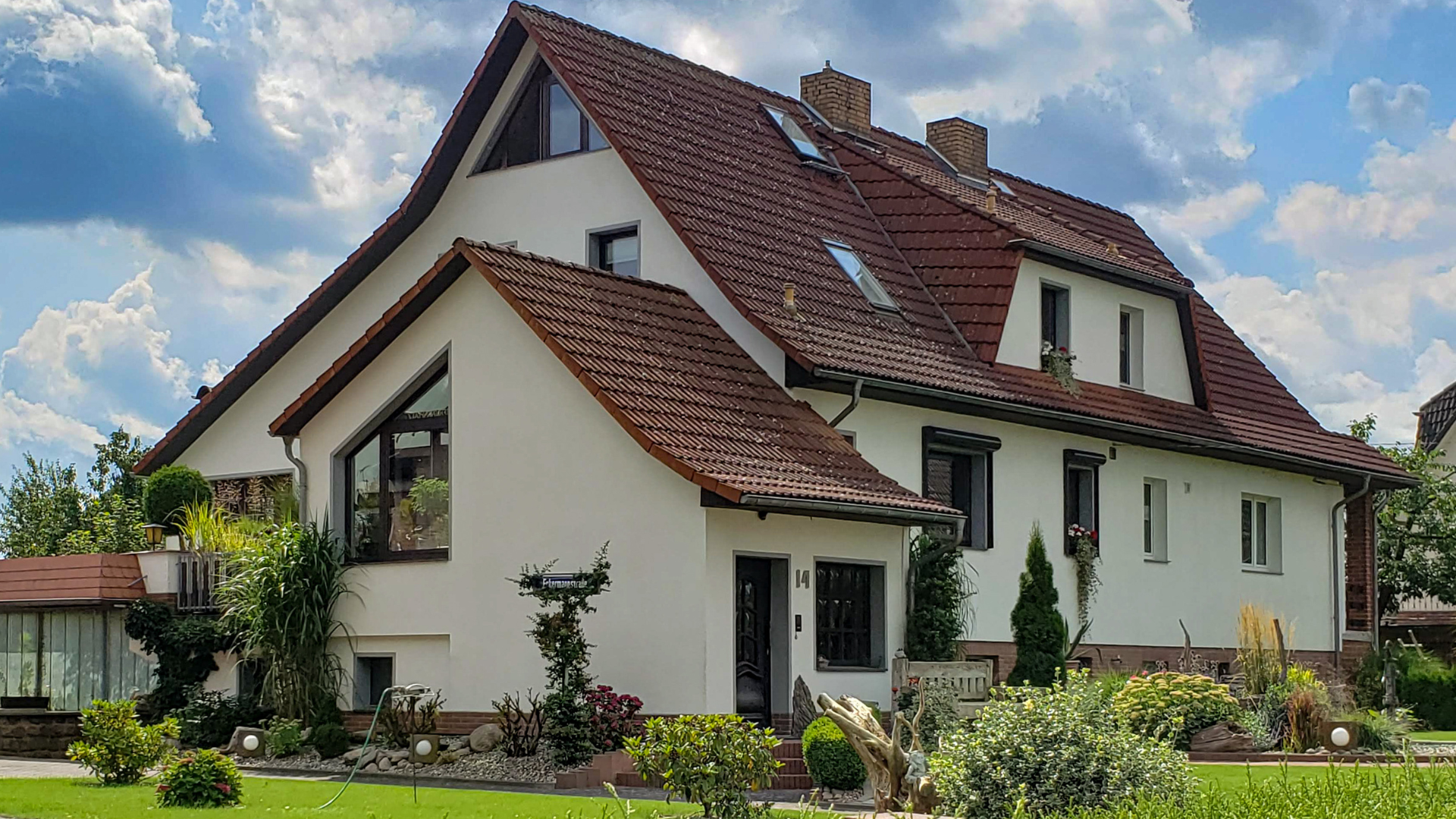 Ein zweistöckiges Haus mit rotem Ziegeldach, weißen Wänden, mehreren Fenstern und einer angebauten Garage, umgeben von einem gepflegten Garten unter einem teilweise bewölkten Himmel.