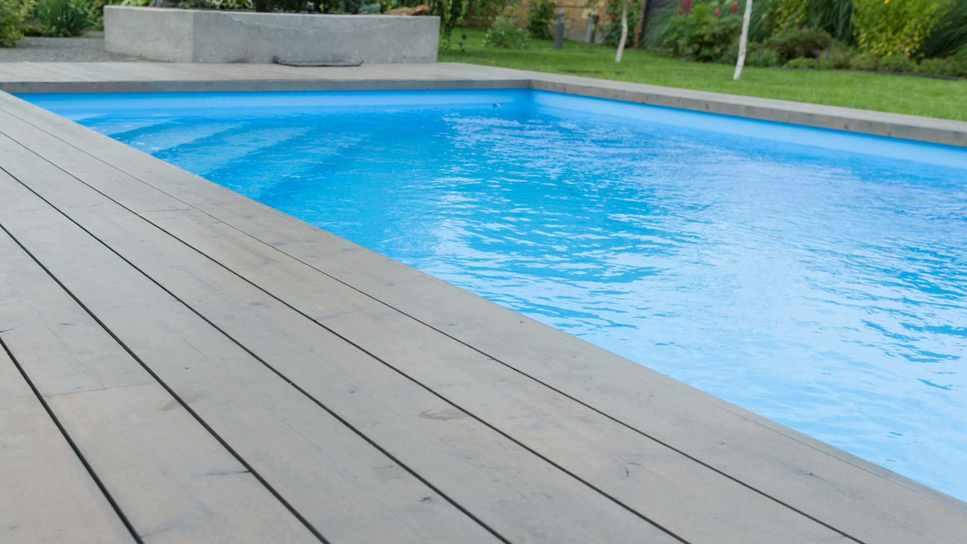 Ein klarer, blauer, rechteckiger Swimmingpool mit Holzterrassen, eingebettet in einen Garten mit Pflanzen im Hintergrund.