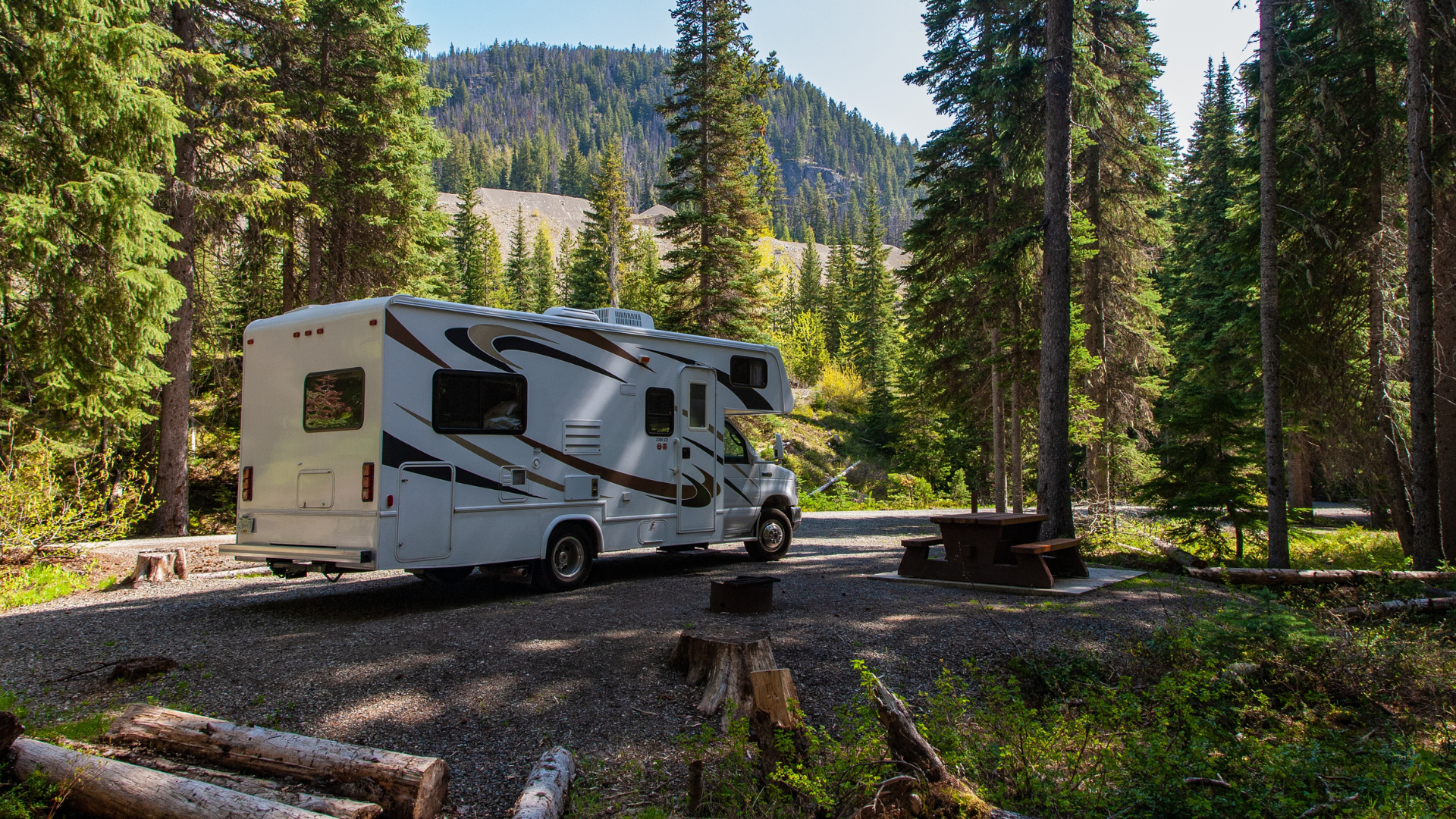Ein weißes Wohnmobil steht auf einem von hohen Bäumen umgebenen Schotterplatz auf einem bewaldeten Campingplatz. In der Nähe stehen ein Picknicktisch und Bänke, im Hintergrund sind Hügel zu sehen.