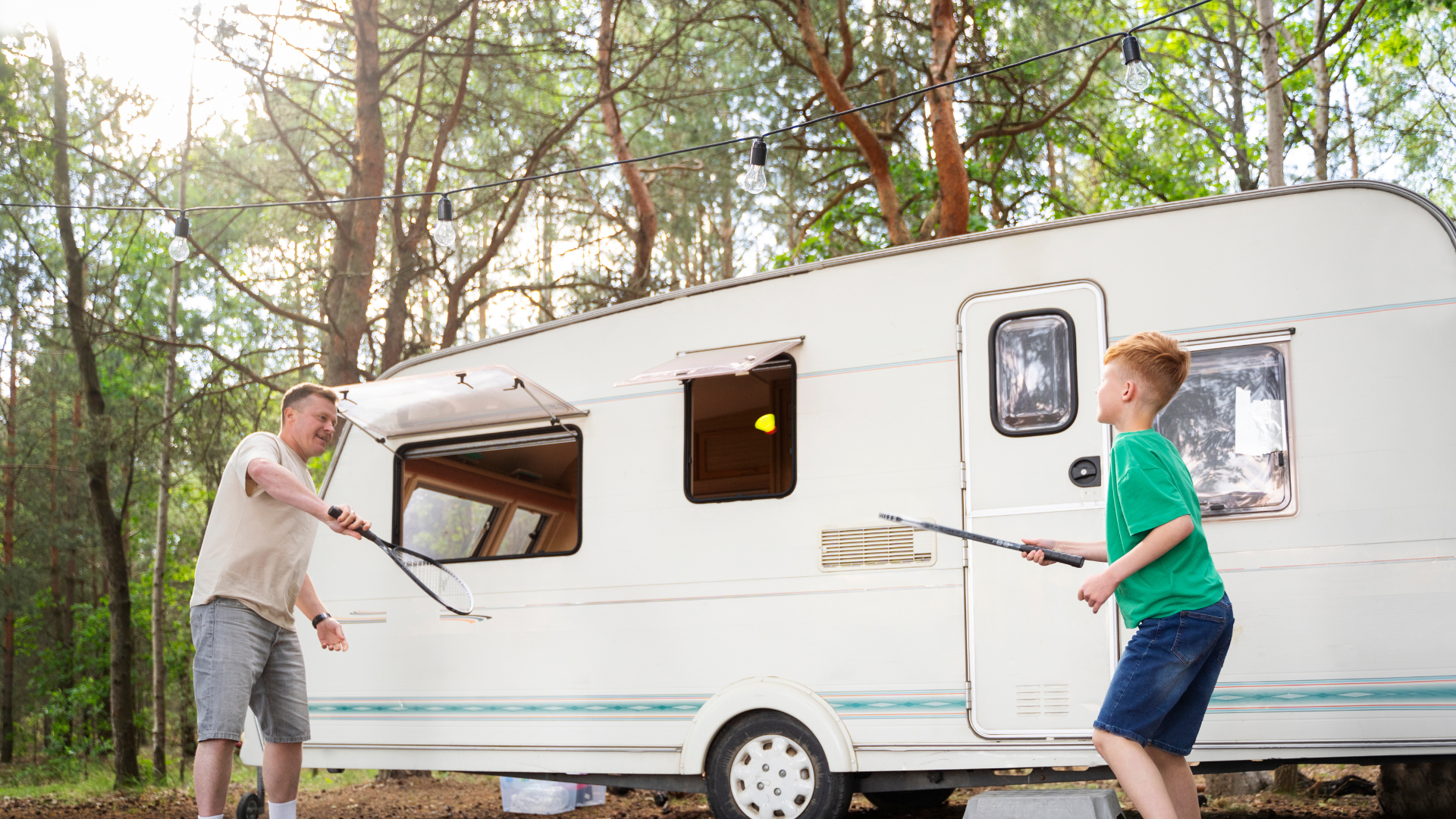 Ein weißes Wohnmobil steht auf einem bewaldeten Campingplatz, umgeben von hohen Kiefern. In der Nähe steht ein Picknicktisch und im Hintergrund ist ein Berg zu sehen.