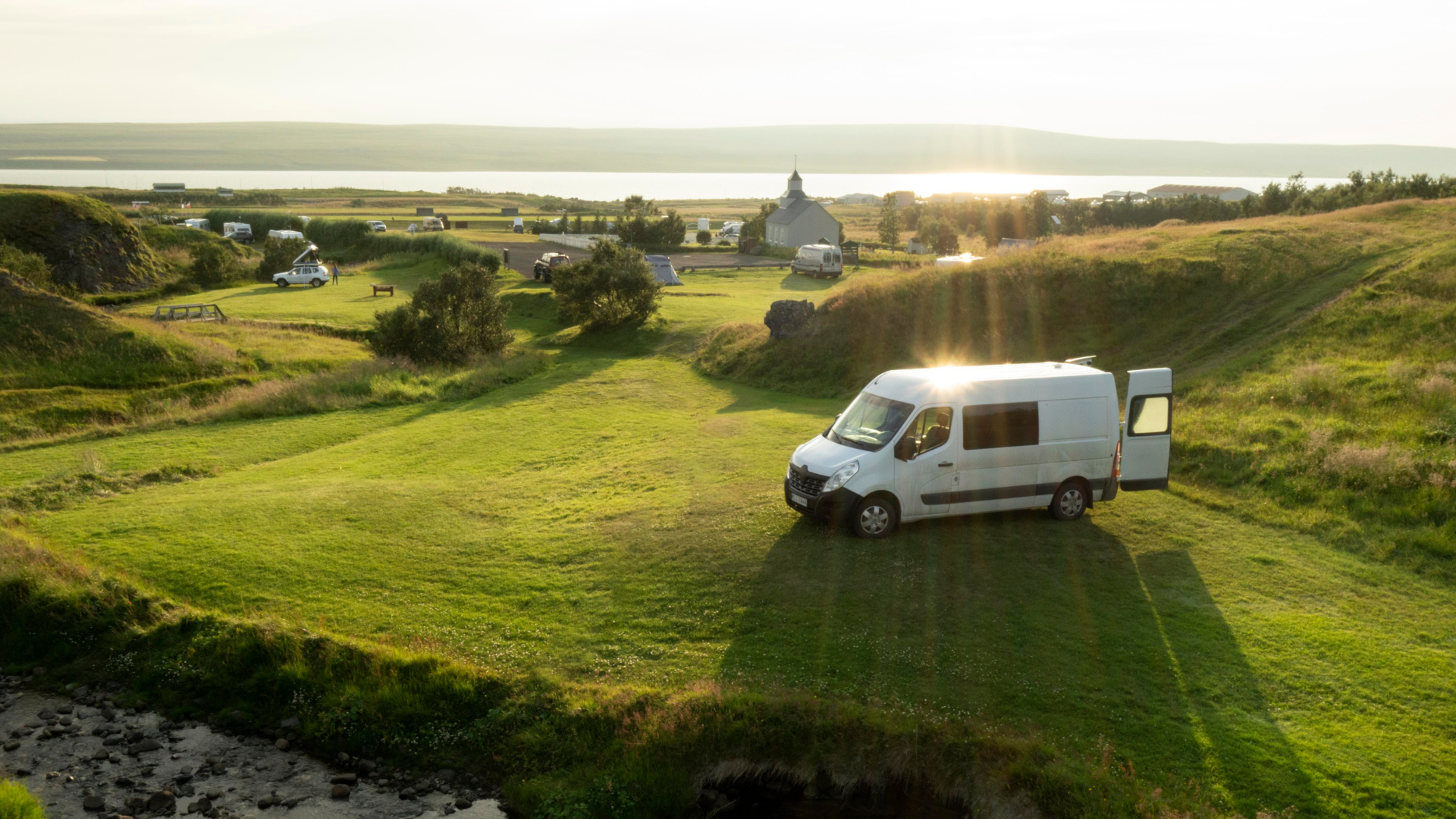 An einem sonnigen Tag steht ein weißes Wohnmobil auf einem bewaldeten Campingplatz mit hohen Bäumen und einem Picknicktisch. Im Hintergrund ist ein Hügel zu sehen.