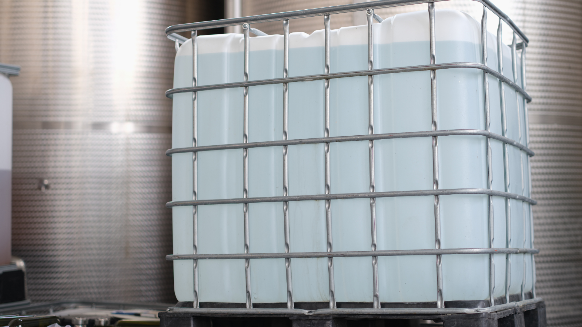 Großer transparenter Tank, gefüllt mit hellblauer Flüssigkeit, mit einem Metallkäfig gesichert, auf einer schwarzen Palette in einer Industrieanlage platziert.