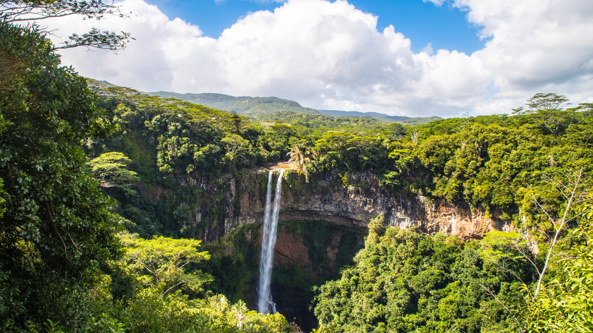 Ein hoher Wasserfall stürzt eine felsige Klippe hinunter, umgeben von dichtem grünen Wald unter einem blauen Himmel mit flauschigen weißen Wolken.