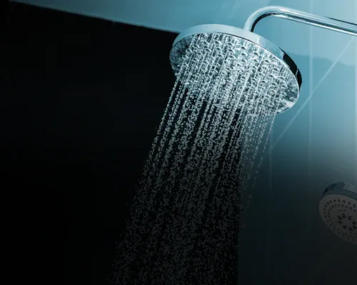 Eine Nahaufnahme eines modernen Duschkopfs, aus dem vor einem dunklen und hellen gefliesten Hintergrund Wasser spritzt.